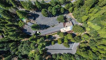 An aerial view of a parking lot surrounded by dense trees, with a building featuring a flat roof and a small patio area.