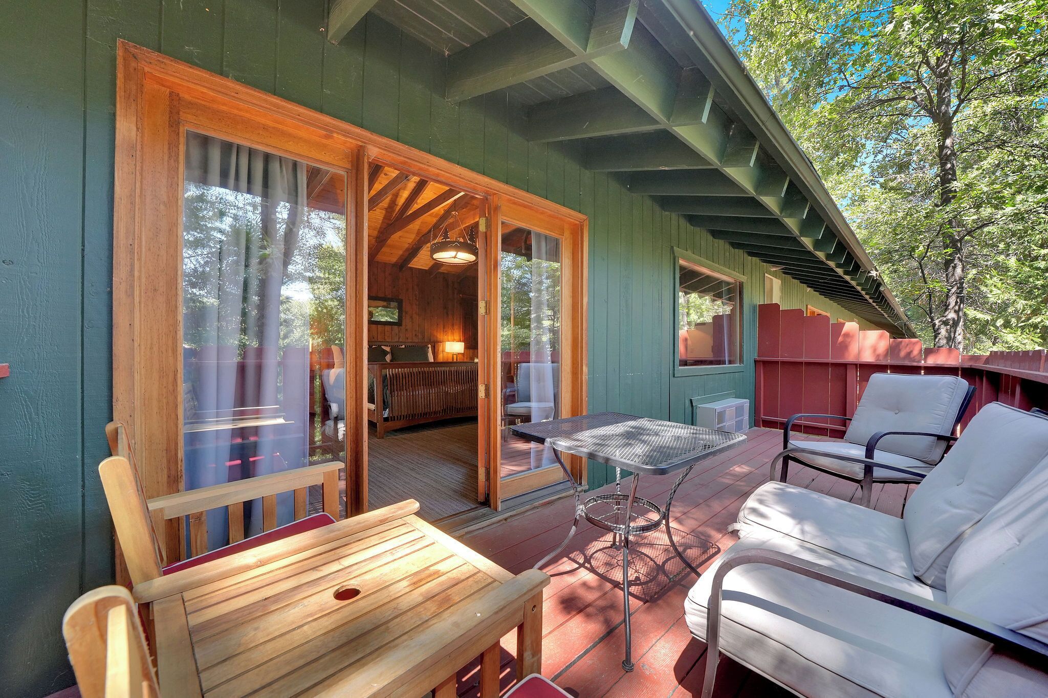 Une terrasse en bois avec une table et des chaises, une porte-fenêtre coulissante donnant sur une pièce avec un canapé et une lampe, et une vue sur les arbres.