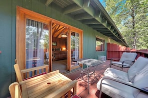 A wooden deck with a table and chairs, a sliding glass door leading to a room with a sofa and a lamp, and a view of trees.