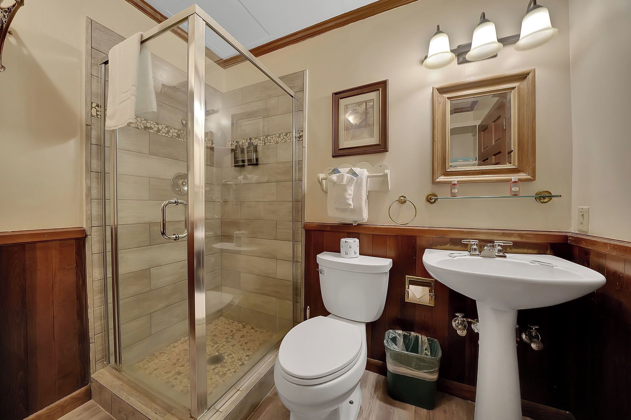 Une salle de bain avec une douche à parois de verre, des toilettes blanches, un lavabo sur colonne et un lambris en bois.