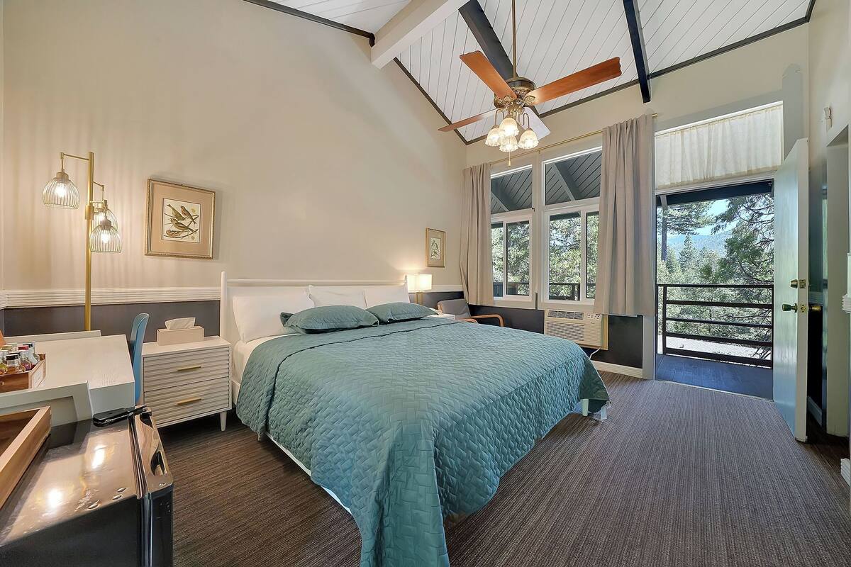 A bedroom with a large bed, a desk, a chair, a ceiling fan, and a window with a view of trees.