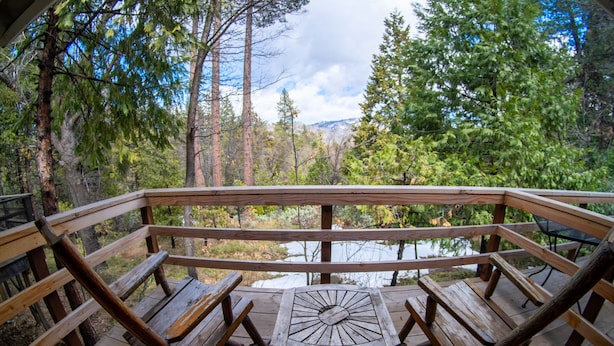 Eine Holzterrasse mit einem Tisch in der Mitte und Bänken, umgeben von hohen Bäumen und Blick auf einen See.