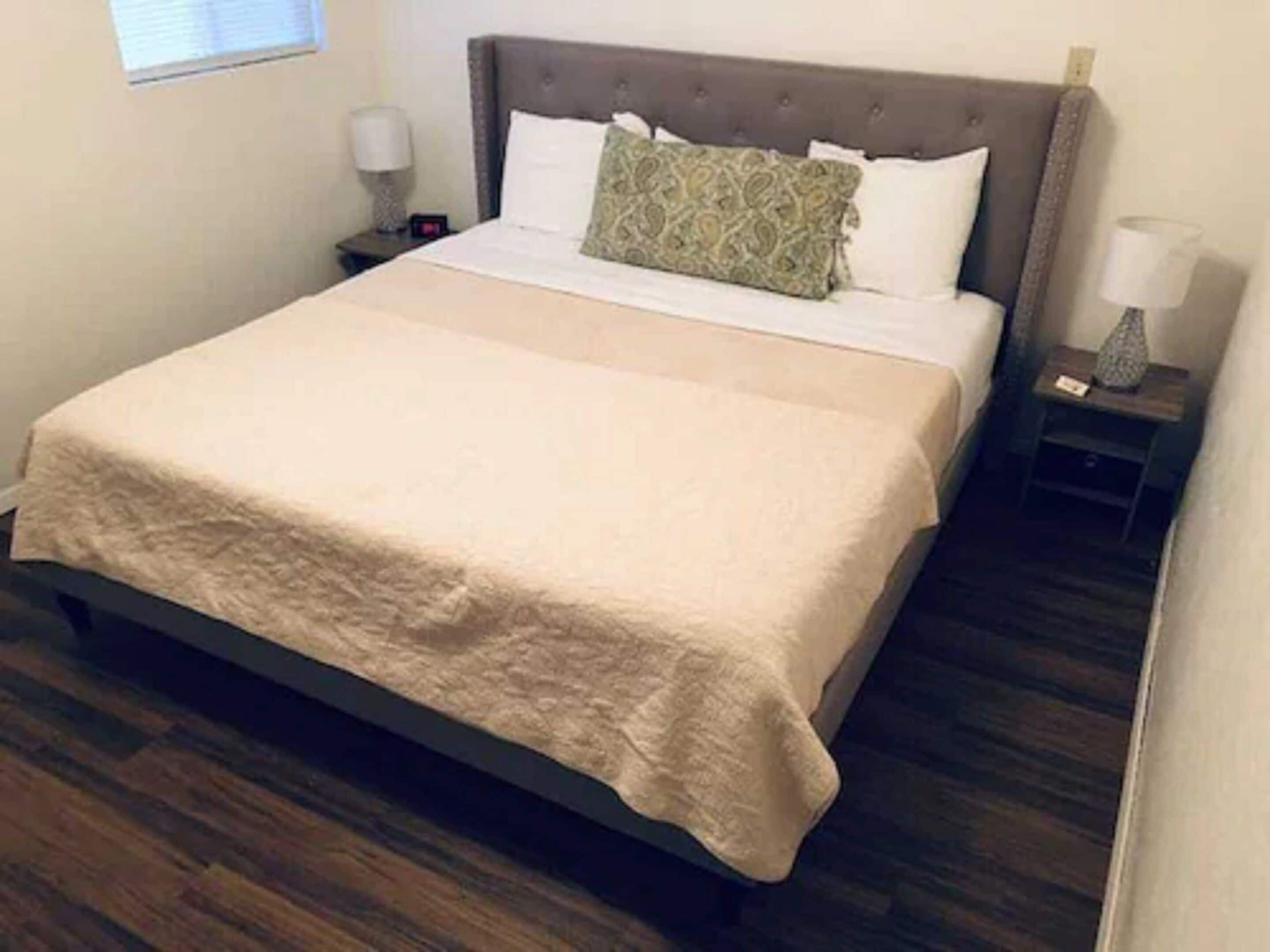 A neatly made bed with a beige bedspread and pillows, a wooden floor, and a nightstand with a lamp.