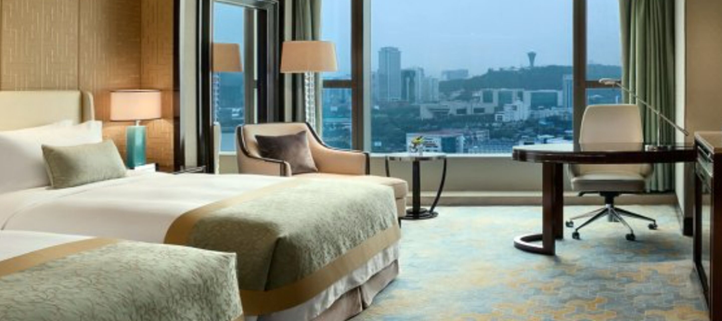 A hotel room with a large bed, a desk, and a chair. There is a view of a cityscape through the window.