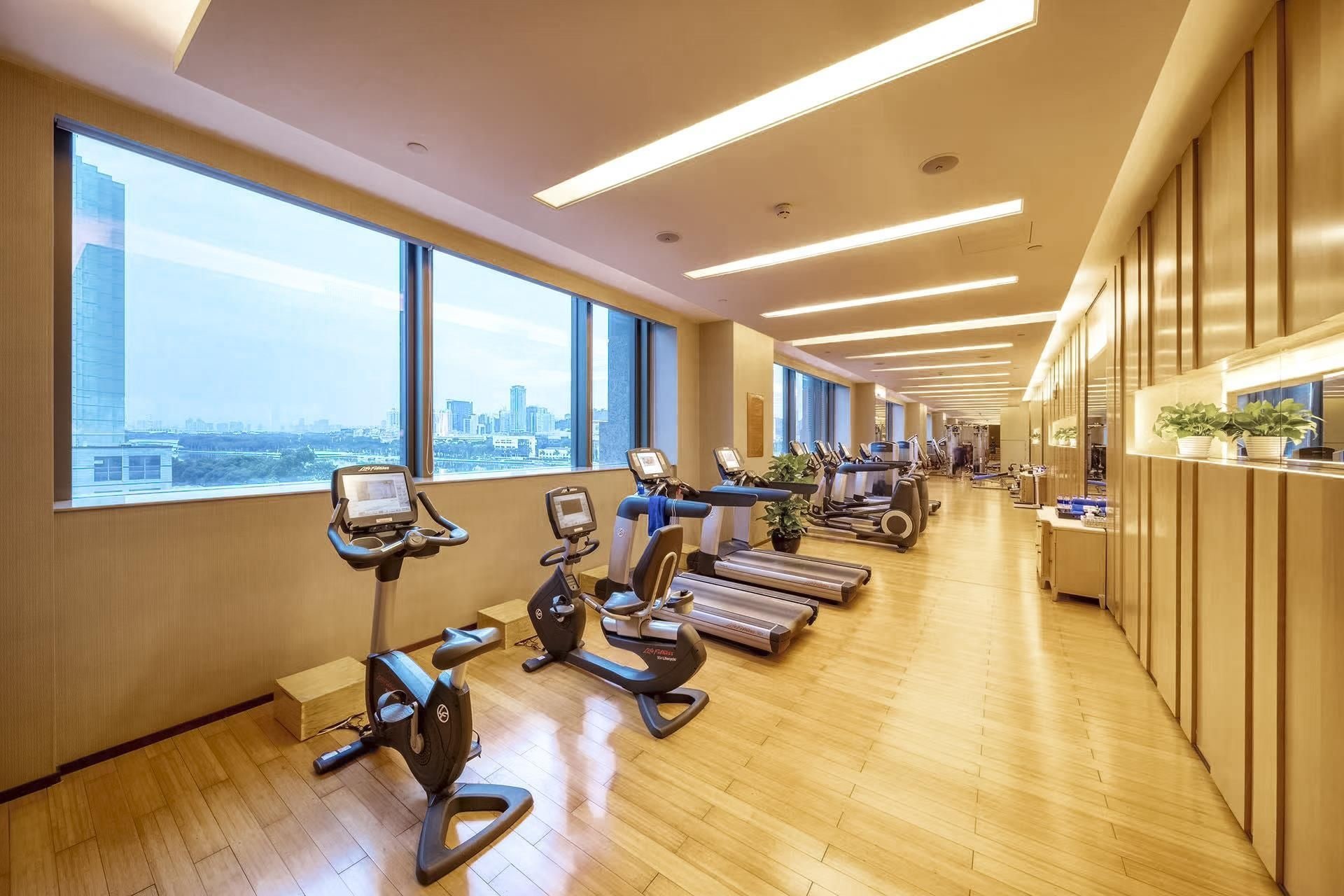 A gym with cardio machines, a wooden floor, large windows, and a city view.