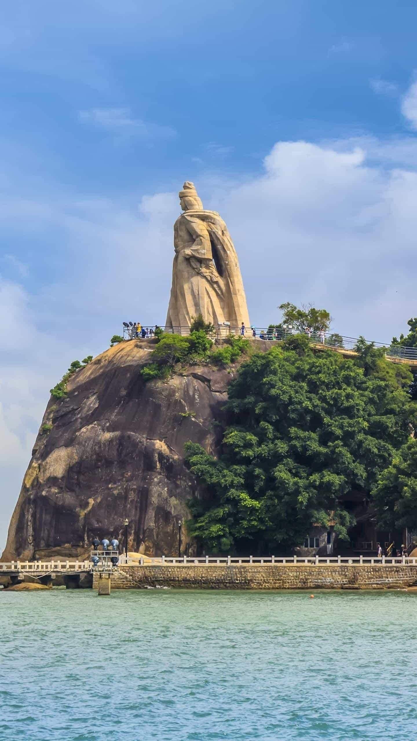 A large rock formation with a statue on top, surrounded by greenery and a body of water.