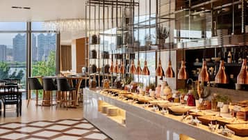 Un moderno vestíbulo de hotel con zona de comedor, un bufé con diferentes opciones de comida y vistas a la ciudad.