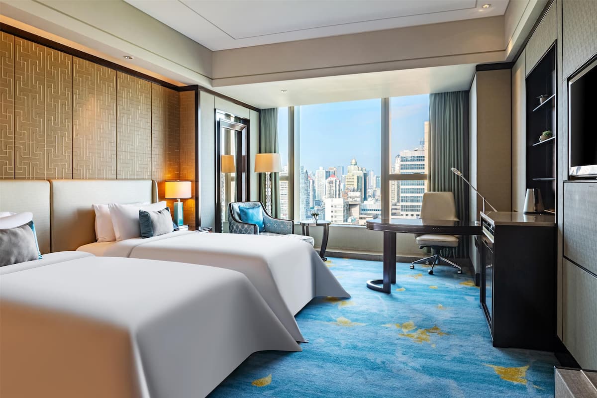 A hotel room with two beds, a desk, a chair, a TV, and a large window with a city view.