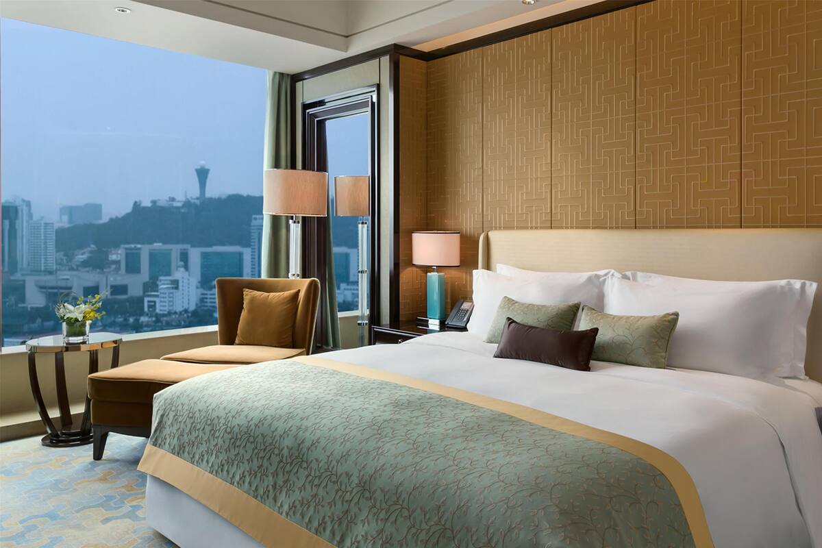 A hotel room with a large bed, a city view, a desk with a lamp, and a chair.