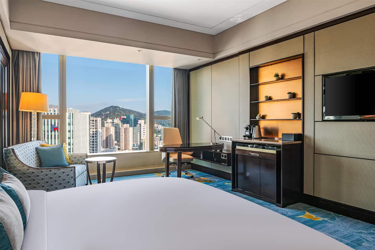 A hotel room with a large bed, a desk, a chair, a TV, and a view of the city.