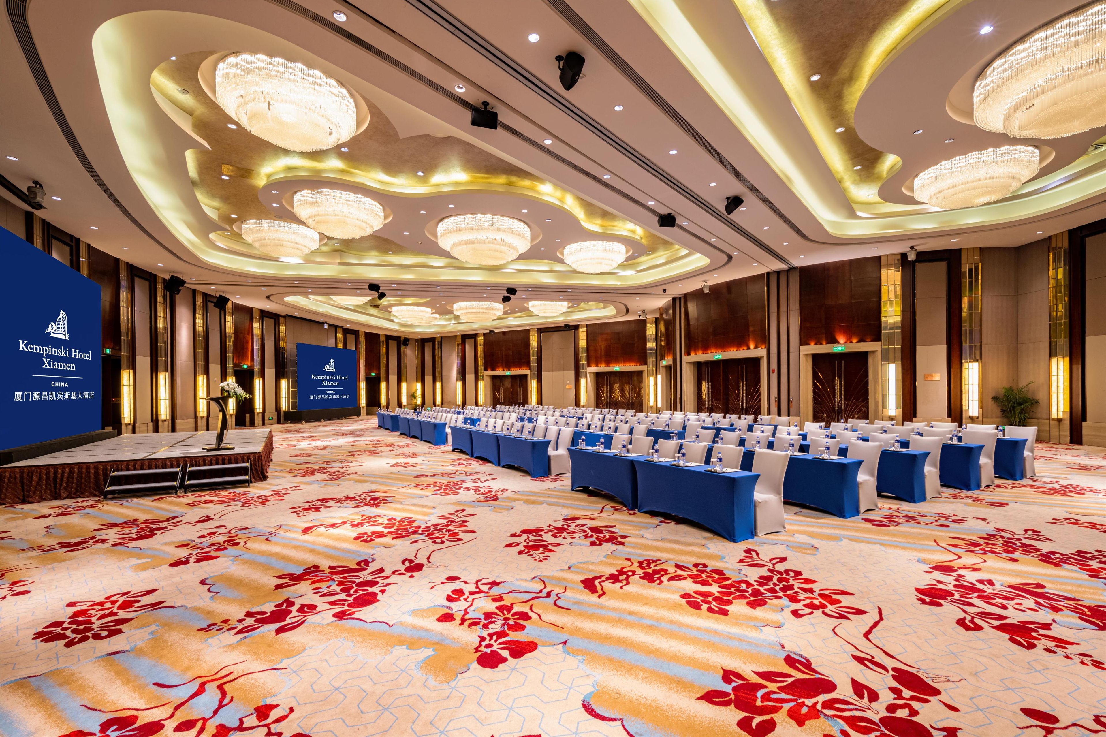 A large conference room with rows of tables and chairs, a stage with a podium, and a floral carpet.