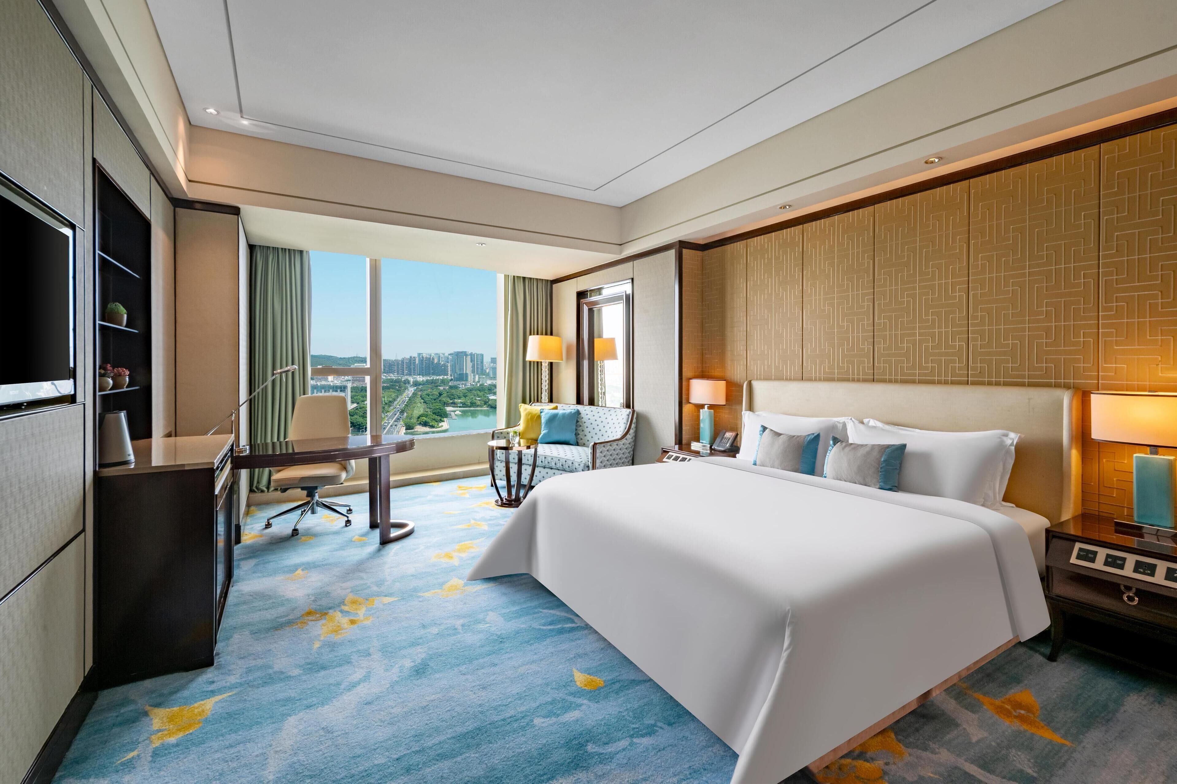 A hotel room with a large bed, a desk, a chair, a TV, and a view of the city.