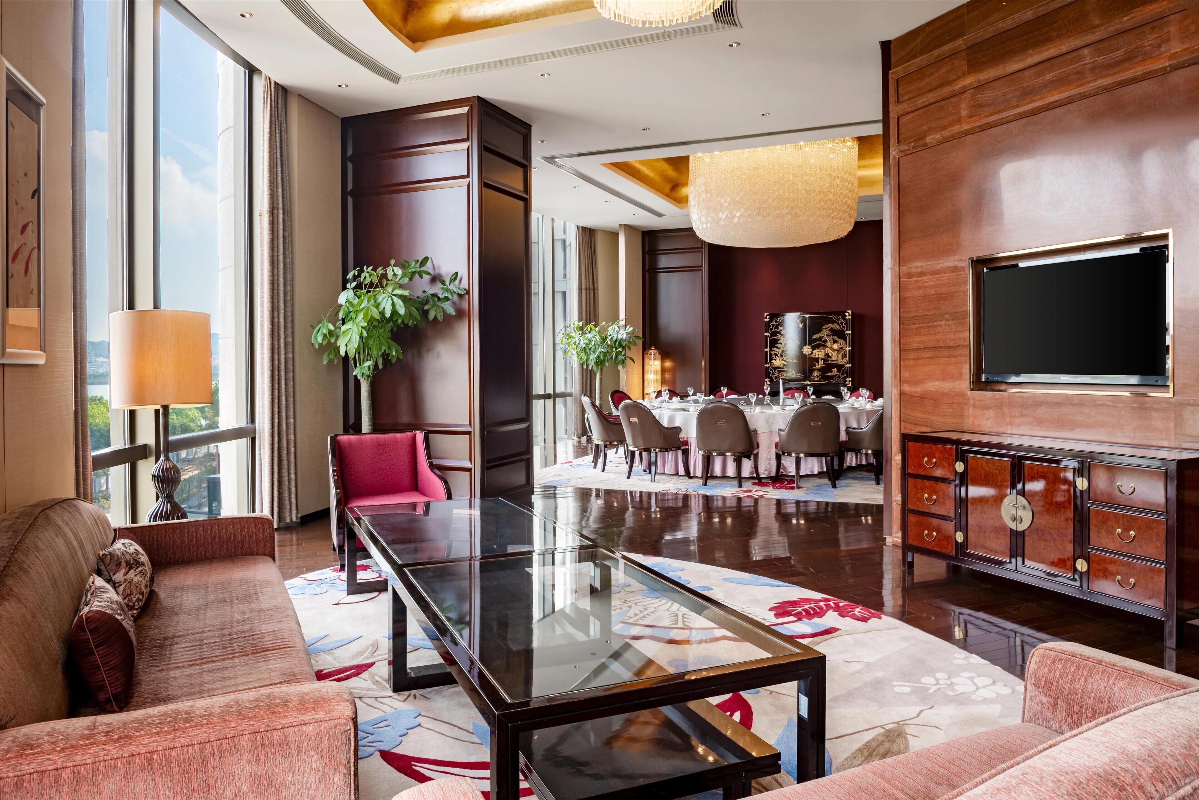 A spacious hotel lobby with a modern glass coffee table, plush seating, a large TV, and a dining area in the background.