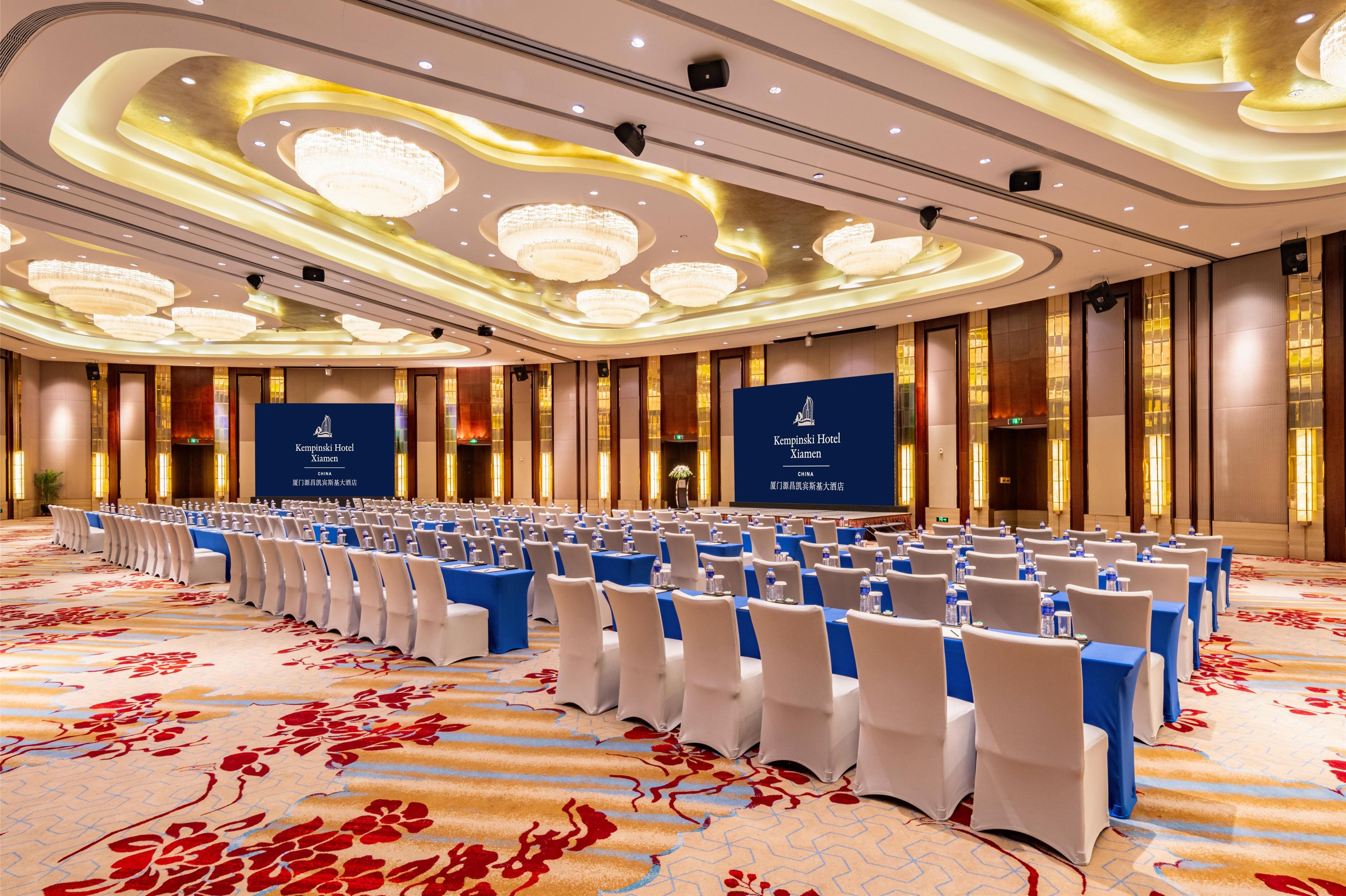 A conference room with rows of tables and chairs, chandeliers, and a carpeted floor.