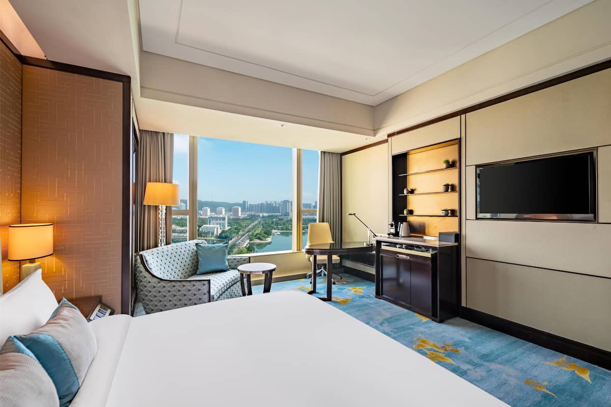 A hotel room with a large bed, a desk, a chair, a sofa, a TV, and a view of the city.