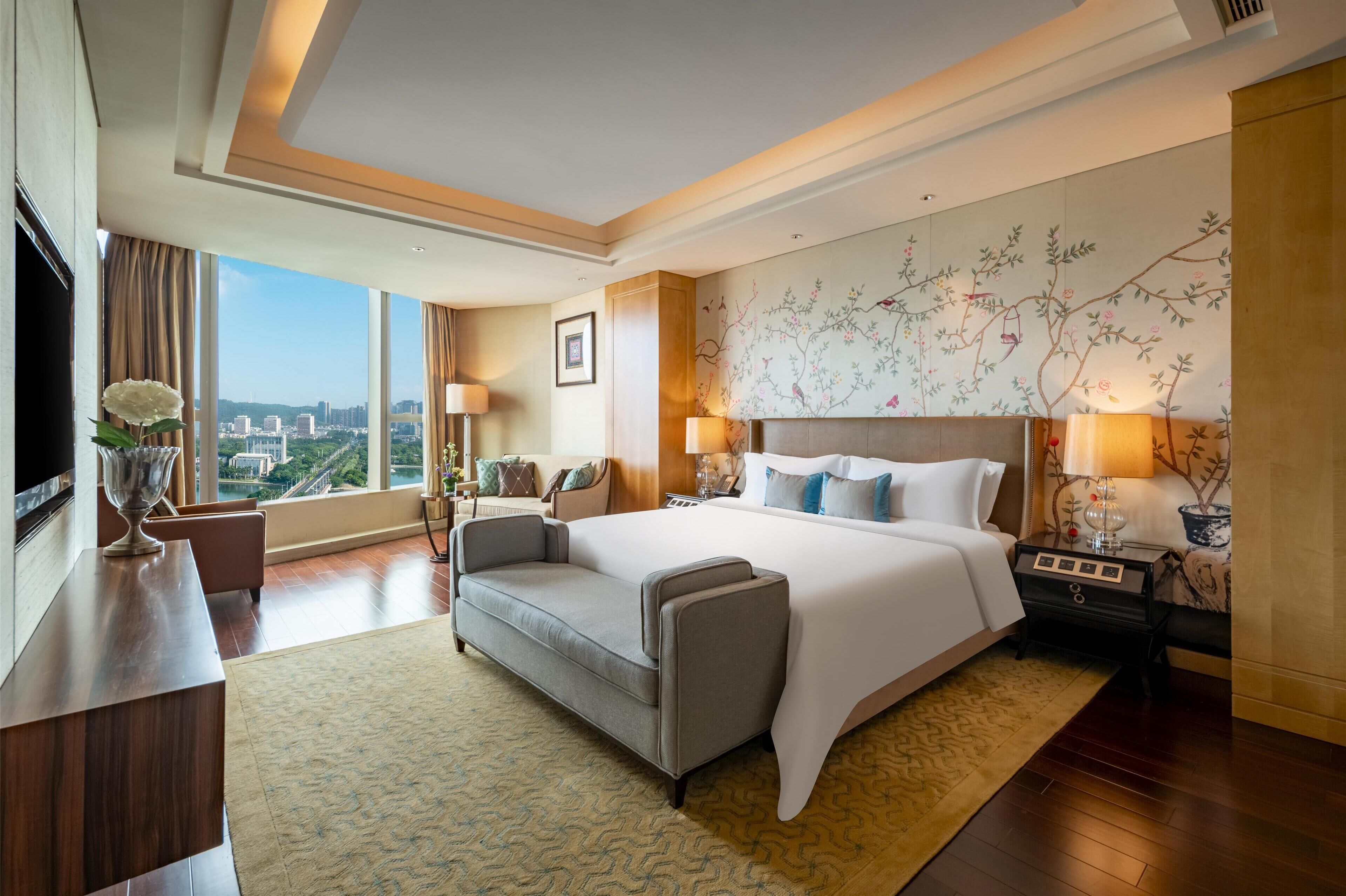 A modern hotel room with a large bed, a sofa, a TV, and a view of the city.