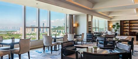 A spacious hotel lobby with large windows offering a city view, comfortable seating areas, and a well-lit interior.
