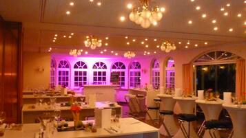 A restaurant with multiple tables and bar stools, a chandelier, and arched windows with purple lighting.