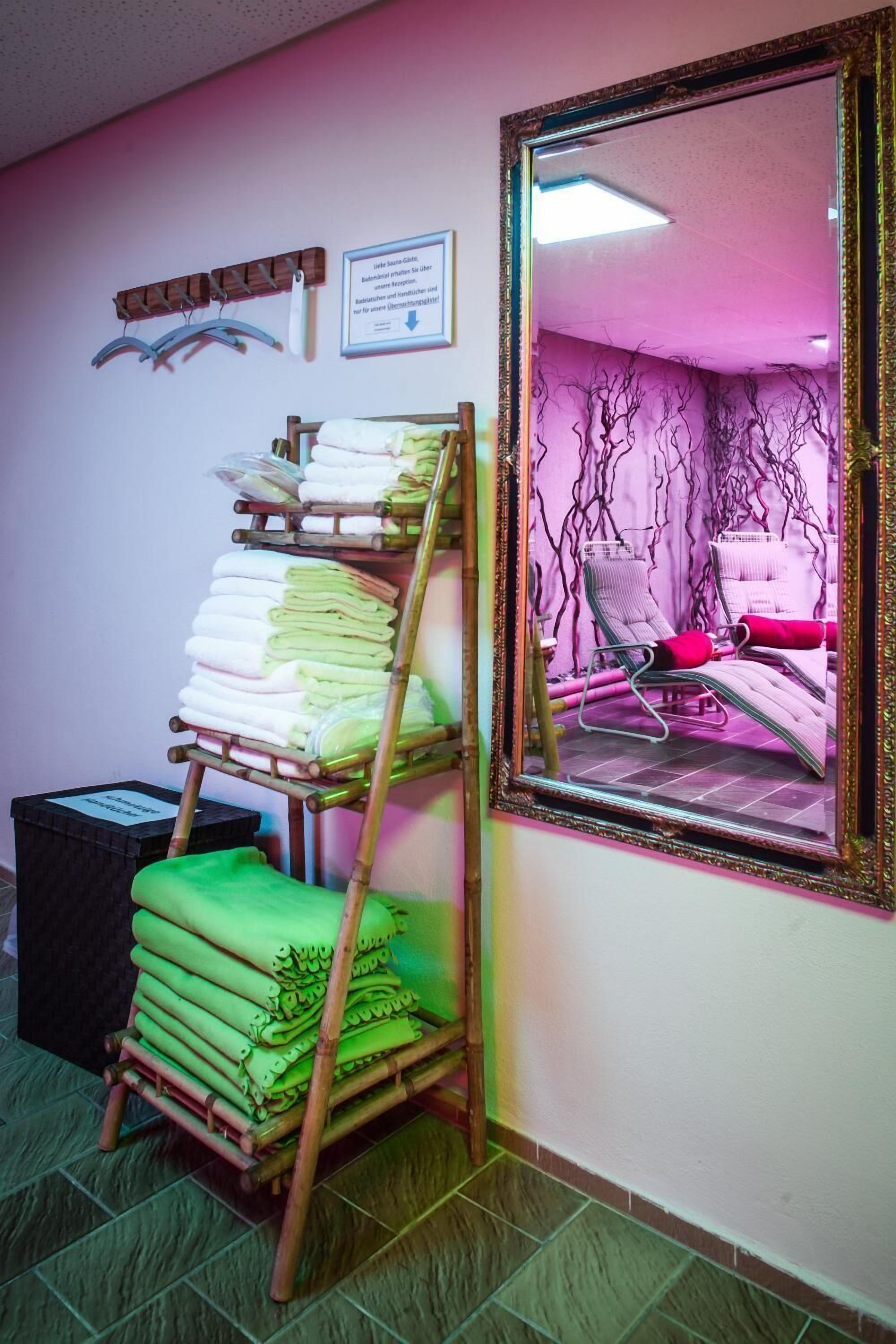 A room with a mirror reflecting a chair and a wall with a decorative vine pattern. A bamboo shelf holds folded towels.