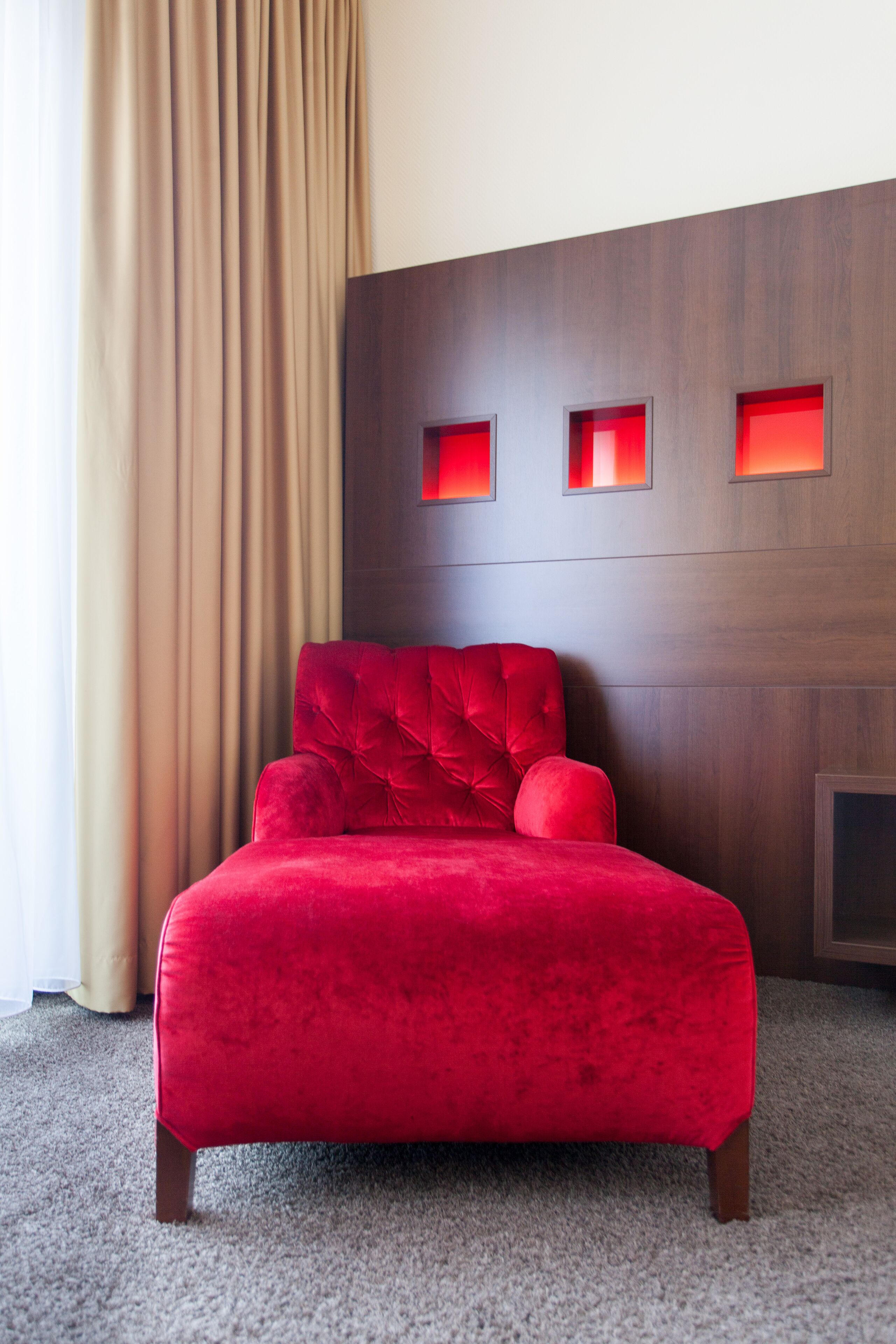 A red velvet bed in a hotel room with a wooden headboard and a beige curtain.