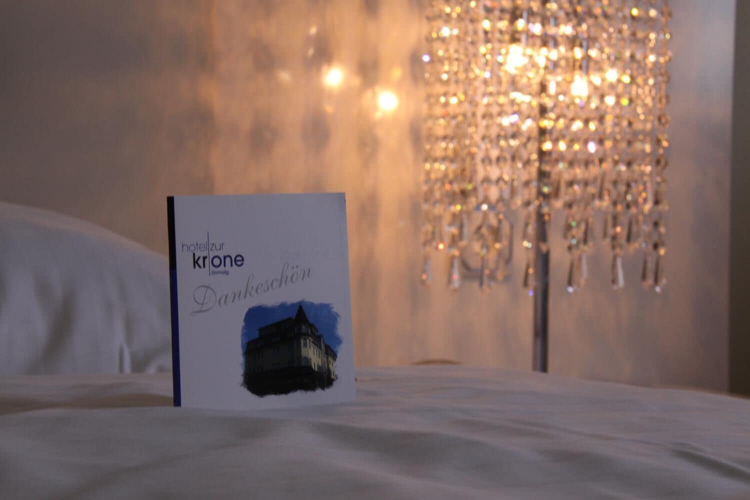 A brochure titled "hotel zur krone" on a bed with a chandelier in the background.