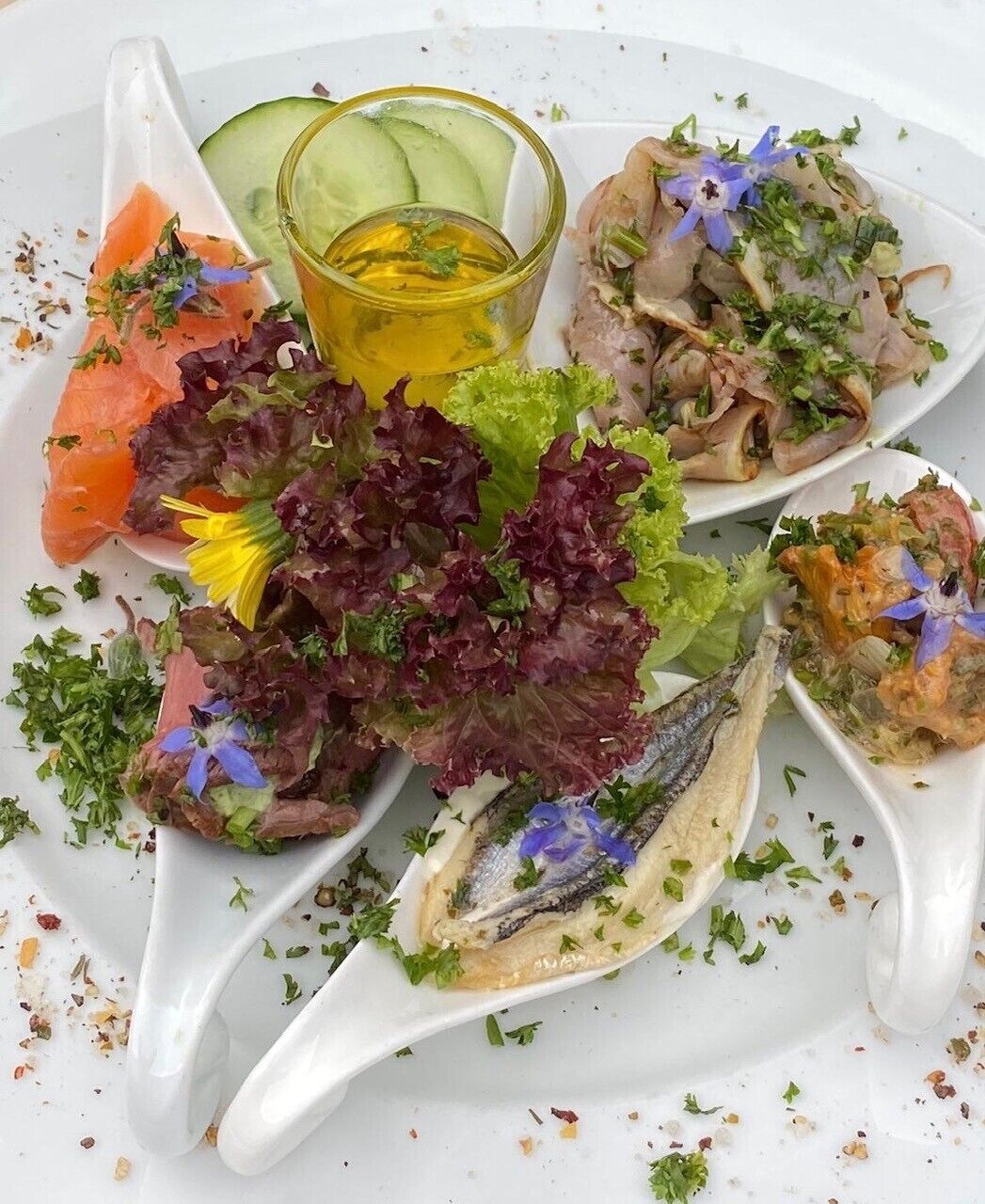A variety of seafood dishes on a white plate, including smoked salmon, a salad with lettuce and herbs, and a fish dish garnished with blue flowers.