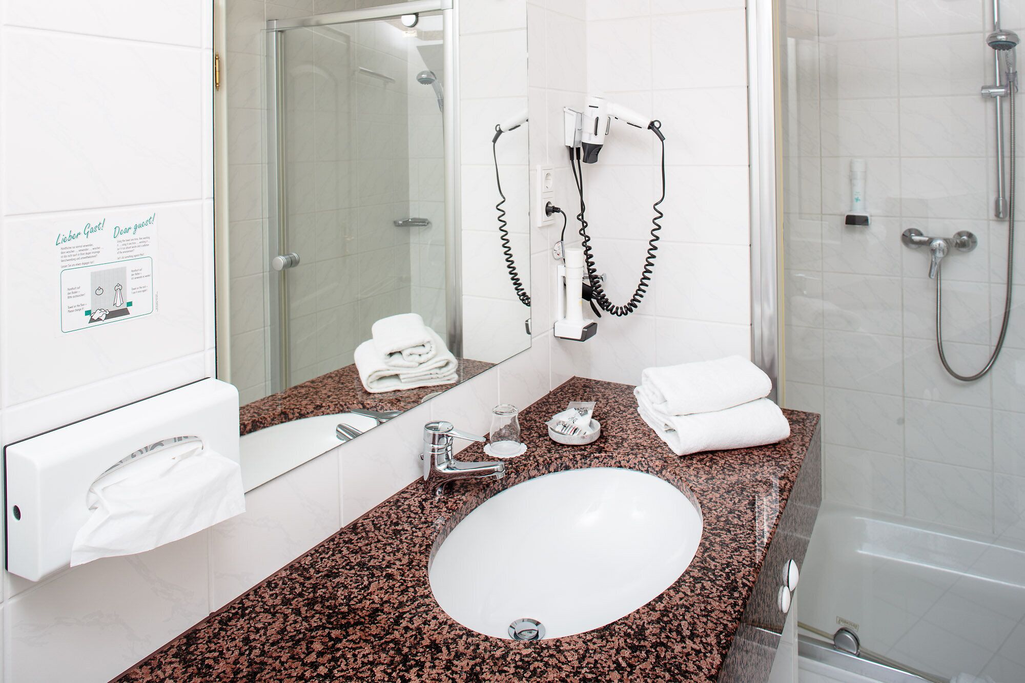 A bathroom with a granite countertop, a white sink, a mirror, a towel rack, a hairdryer, and a shower.