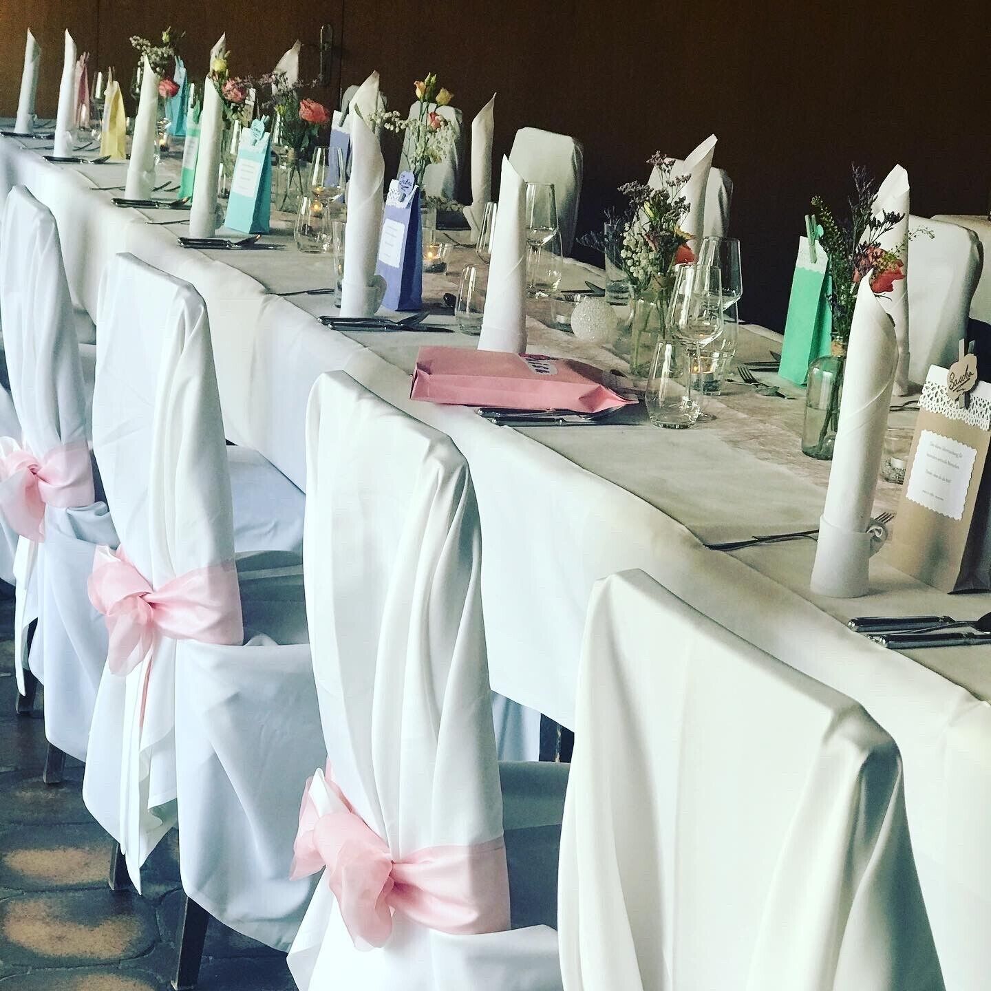 Une longue table de banquet, recouverte de nappes blanches, avec des chaises habillées d’un tissu blanc agrémenté de rubans roses.