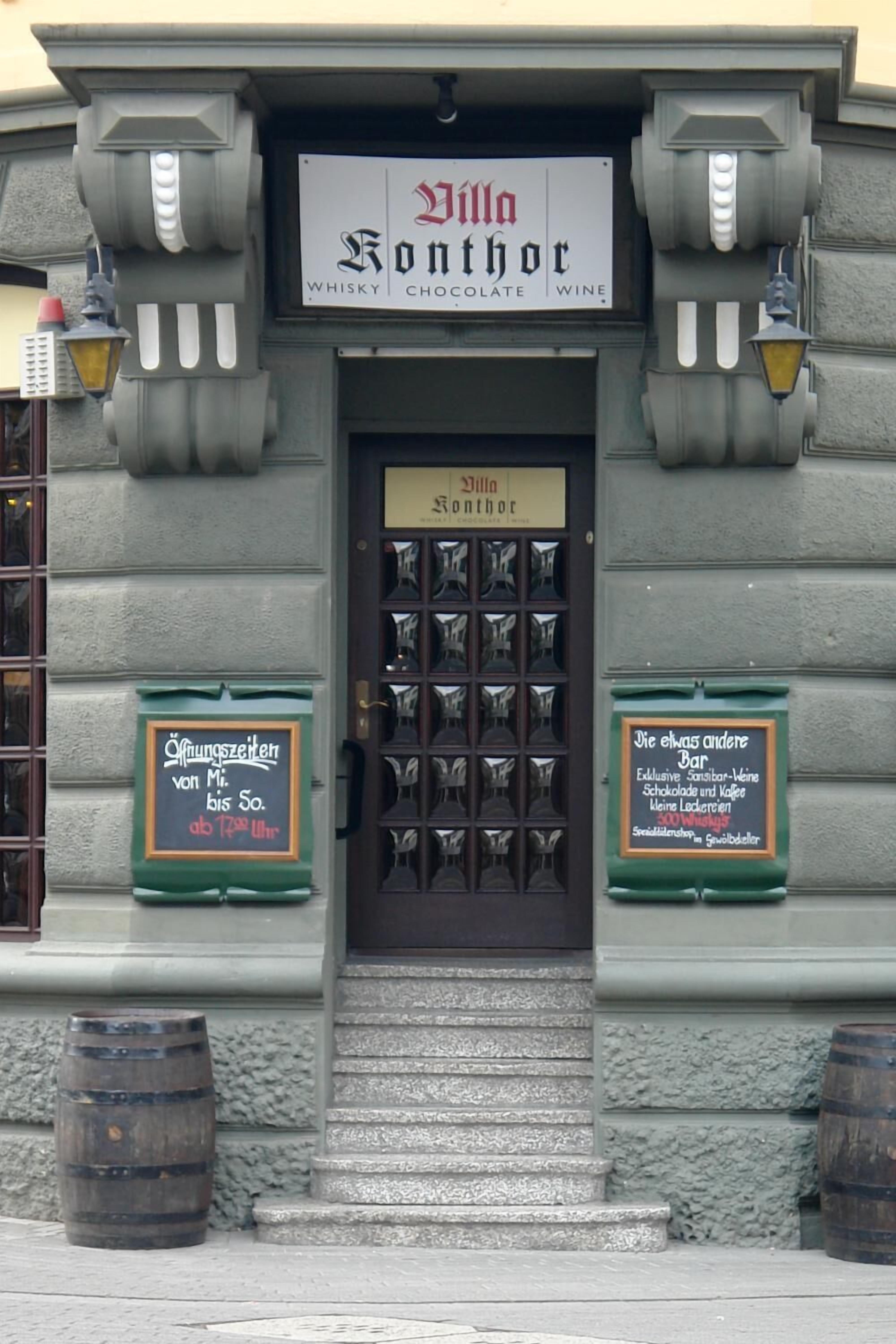 A building with a sign reading "Villa Konthor" and a door labeled "3. Villa Konthor." There are two green signs on either side of the door, one with a price and the other with a description of the bar.
