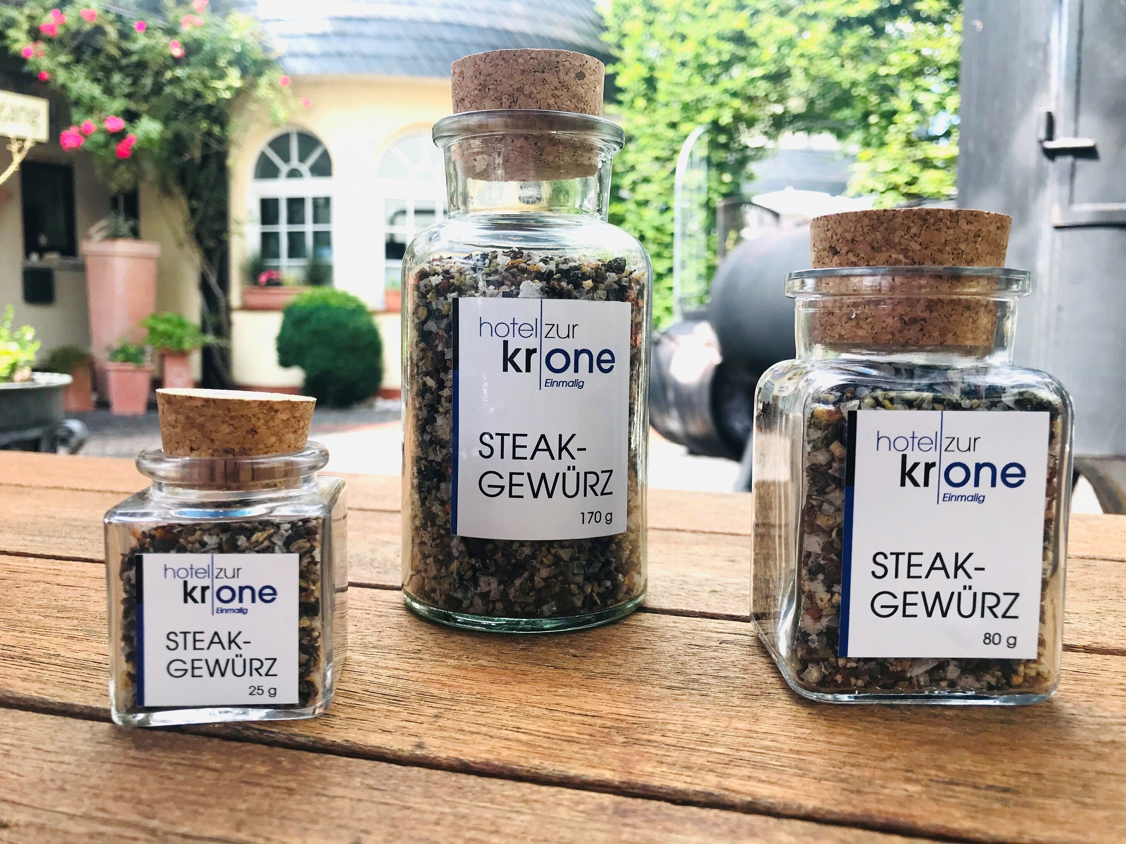 Tres frascos de vidrio con la etiqueta 'Hotel zur Krone' que contienen especias para carne, sobre una mesa de madera al aire libre.