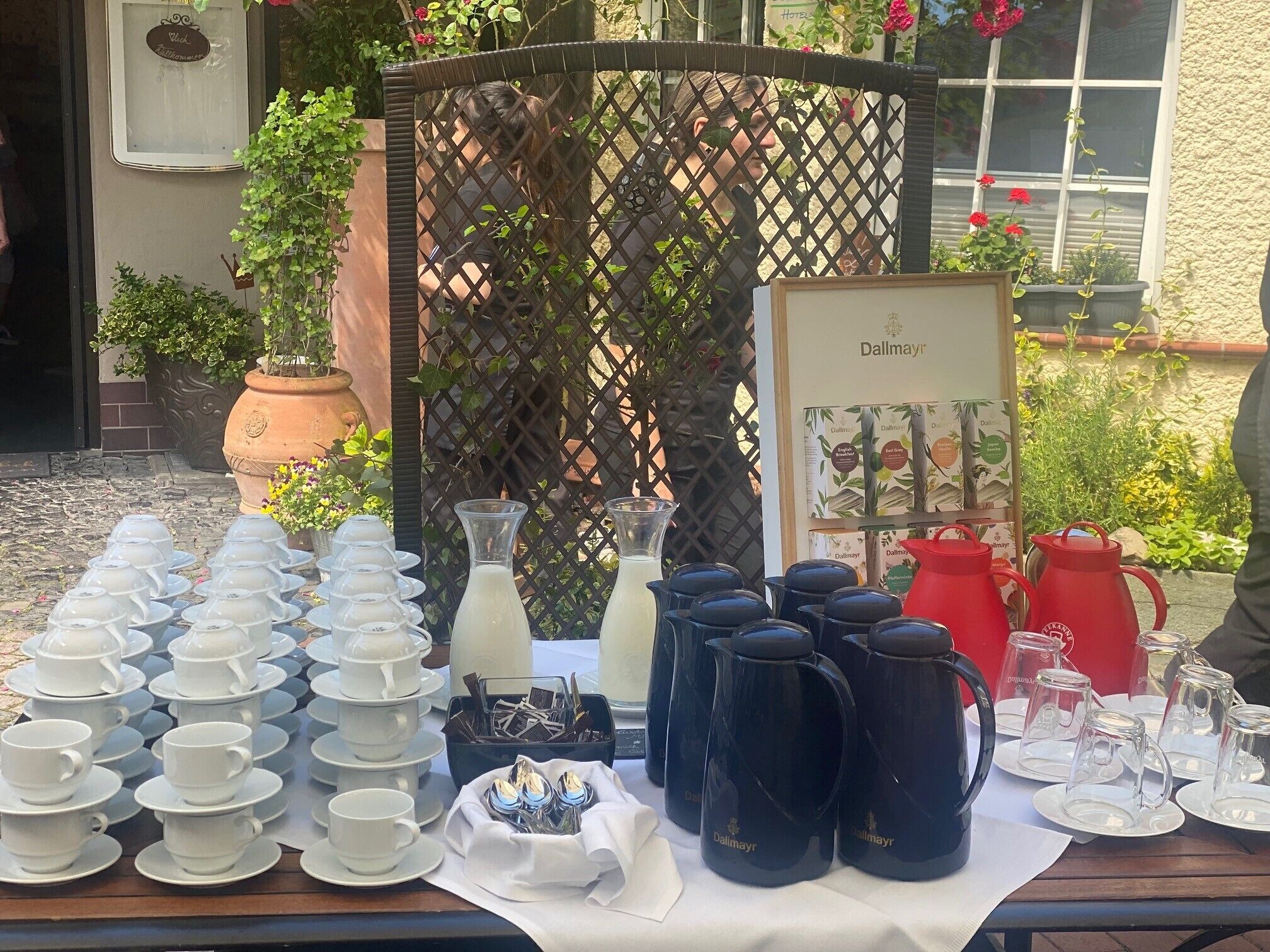 A table set with tea service items, including teapots, cups, and saucers, in an outdoor setting with a lattice backdrop and potted plants.