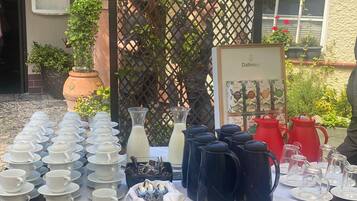 A table set with tea service items, including teapots, cups, and saucers, in an outdoor setting with a lattice backdrop and potted plants.