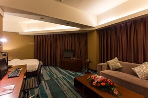 Quarto de hotel com cama, escrivaninha com computador, televisão, sofá e mesa de centro com flores.
