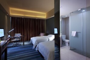 Quarto de hotel com duas camas, uma escrivaninha com computador, uma cadeira, luminária de mesa e uma janela grande com cortinas.