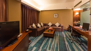 A hotel room with a TV, sofa set, coffee table, desk with computer, and a patterned carpet.