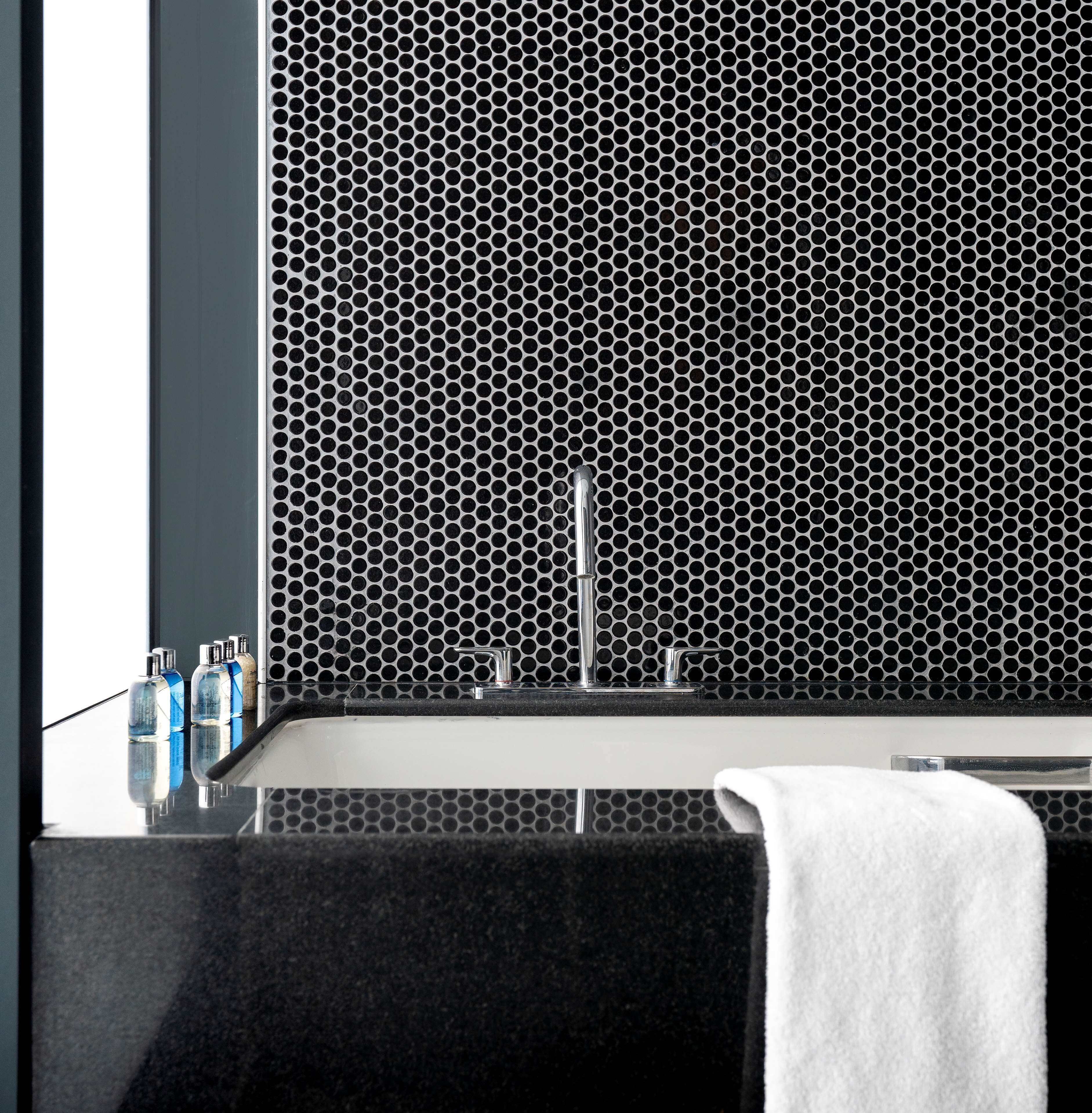 Un baño con pared de azulejos hexagonales blanco y negro, un lavamanos y un espejo. Hay tres botellas de jabón líquido en la pared.