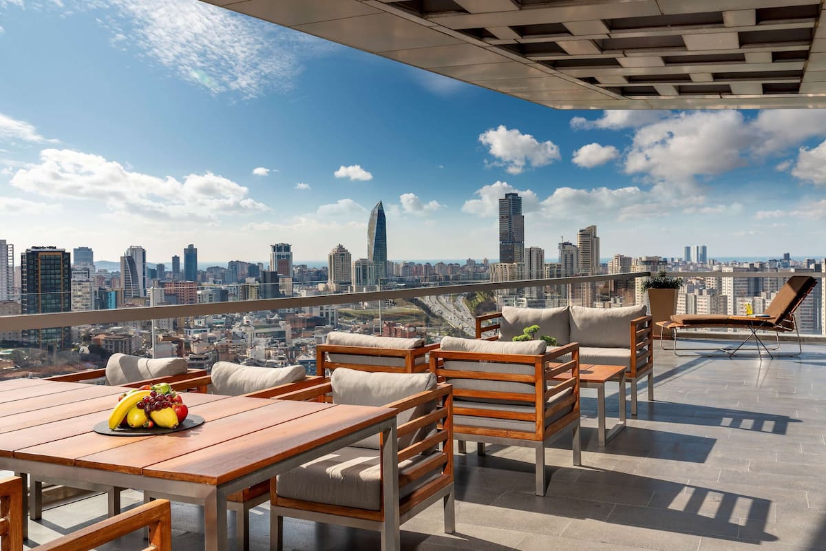 A rooftop terrace with wooden furniture, a table with fruit, and a city skyline view.