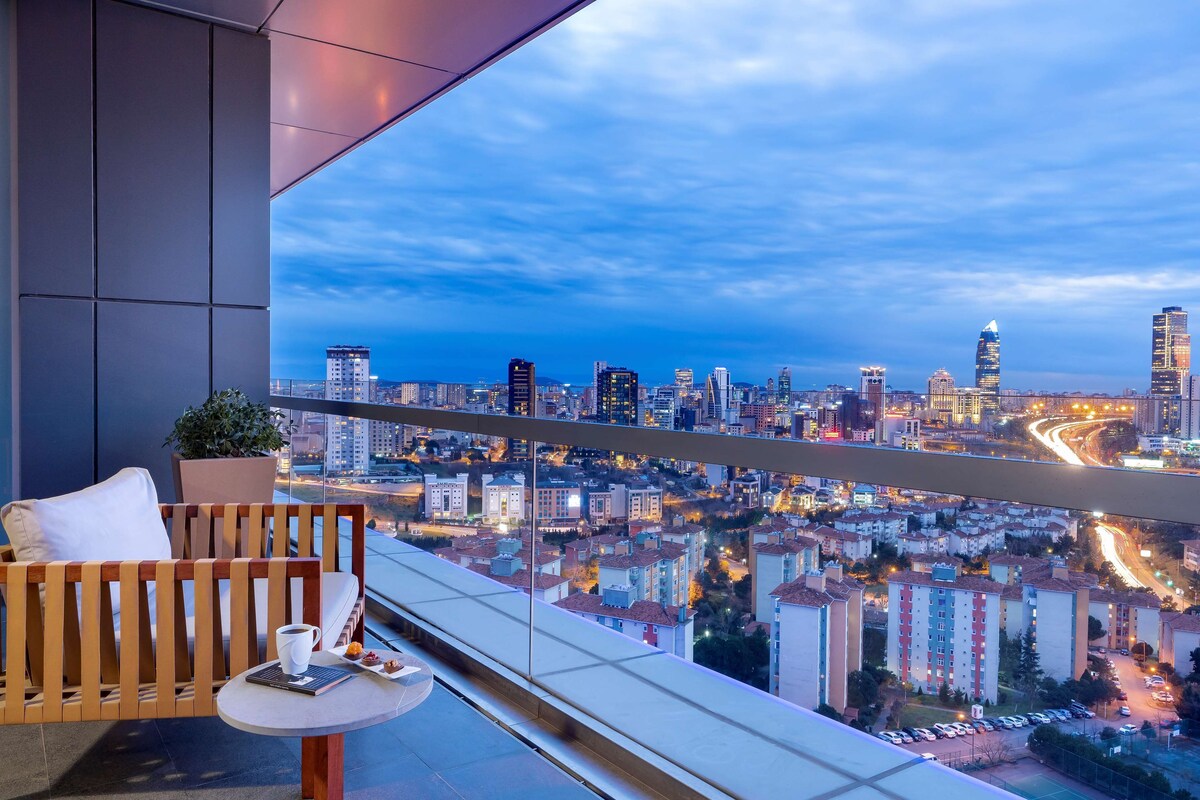 Un balcon avec vue sur la ville, une chaise en bois, une petite table avec une tasse et une assiette, et une plante en pot.