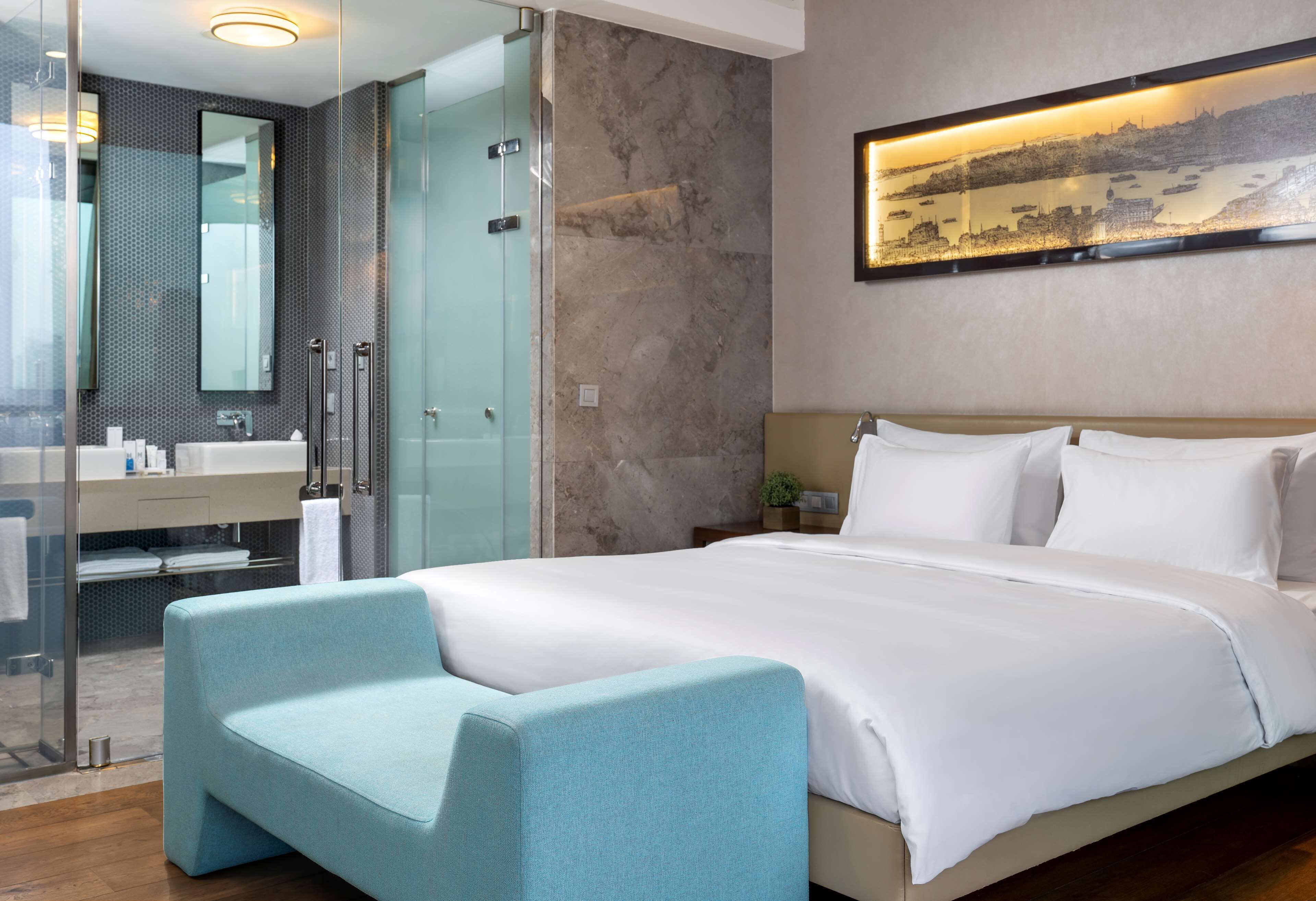 Una habitación de hotel moderna con una cama grande, zona de estar, ducha de vidrio, baño con lavabo y espejo, y un cuadro de paisaje urbano en la pared.