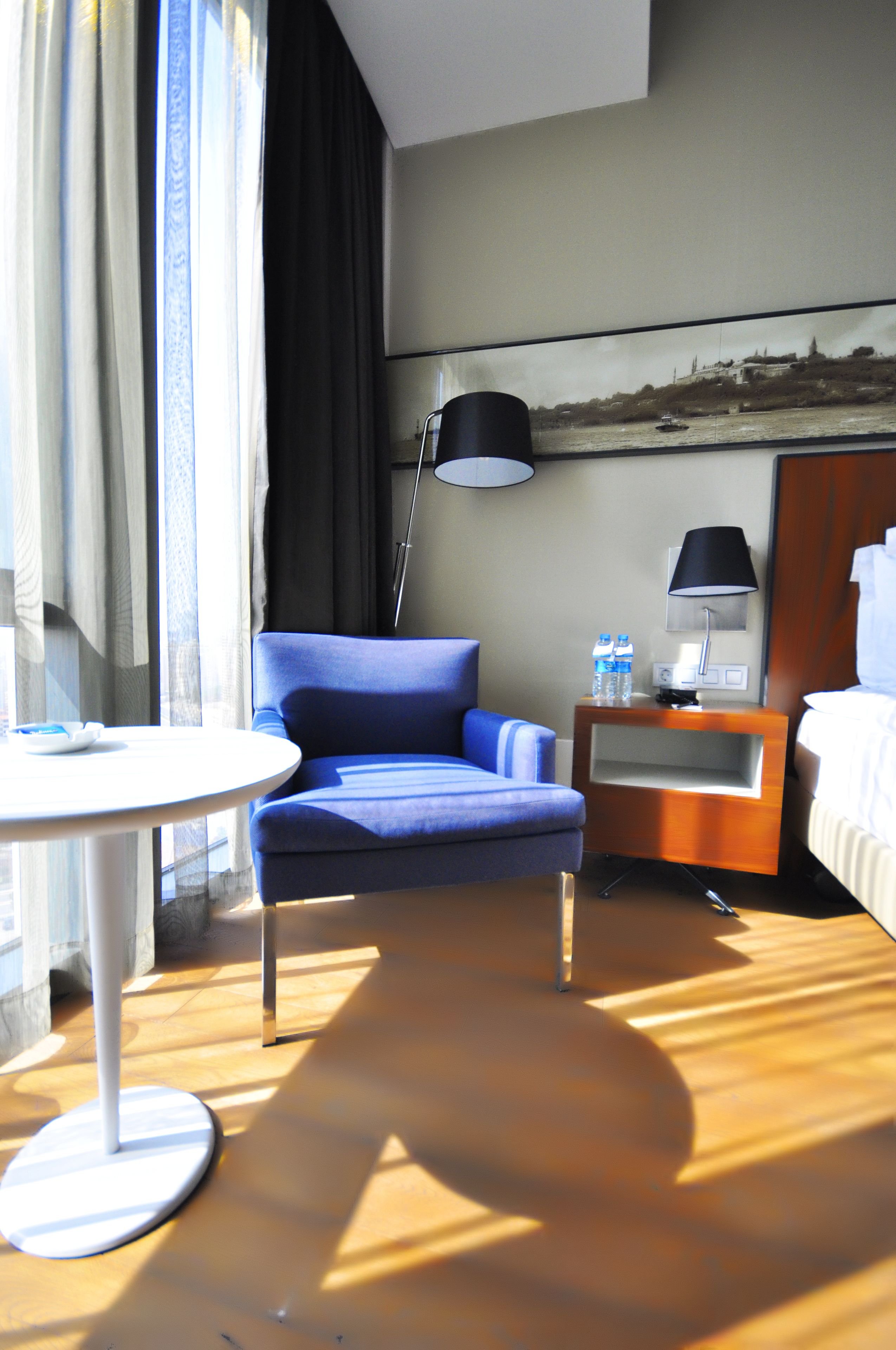 Ein Hotelzimmer mit blauem Sessel, kleinem runden Tisch, Nachttischlampe, Wasserflasche und einem Bett.