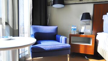Ein Hotelzimmer mit blauem Sessel, kleinem runden Tisch, Nachttischlampe, Wasserflasche und einem Bett.
