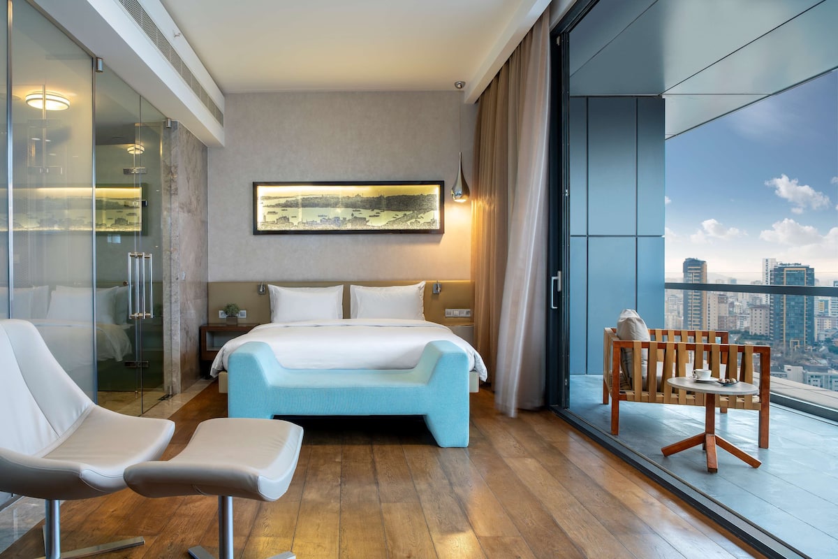 A modern hotel room with a large bed, a seating area with a table, and a city view through large windows.