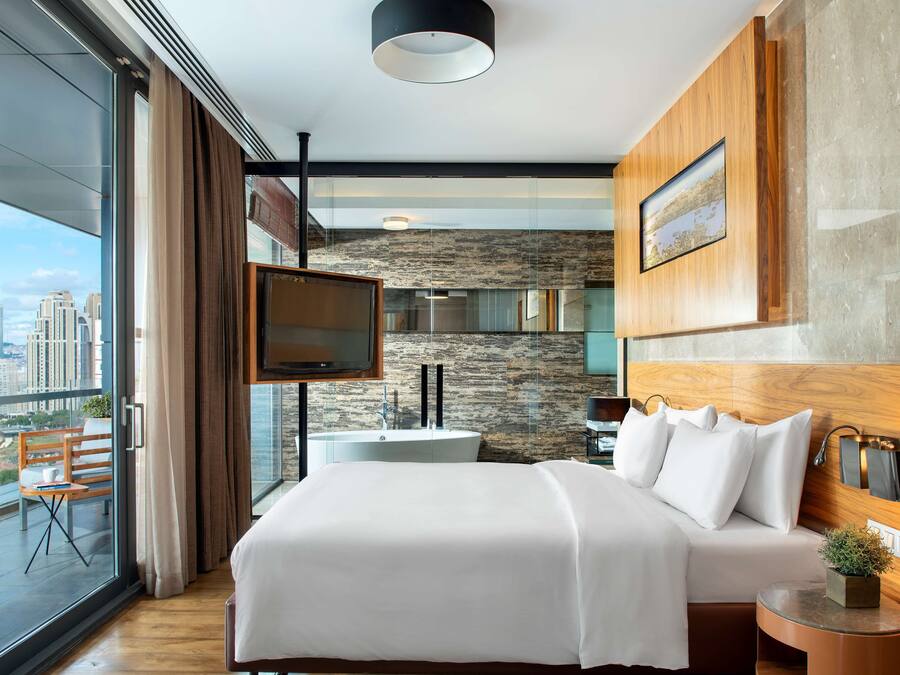 Ein modernes Hotelzimmer mit einem großen Bett, einem an der Wand montierten Fernseher, einem Balkon mit Blick auf die Stadt und einer Wand mit Steinoptik.