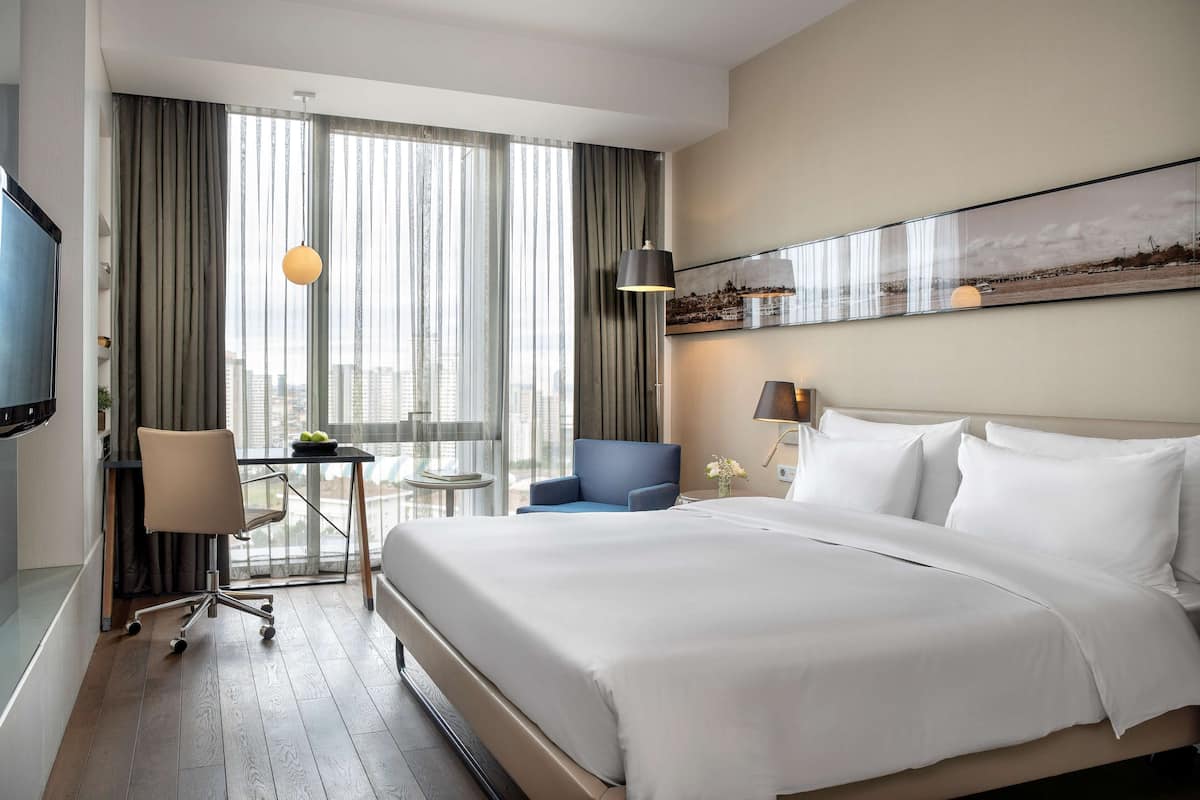 A hotel room with a large bed, a desk with a chair, a television, and a view of the city through large windows.