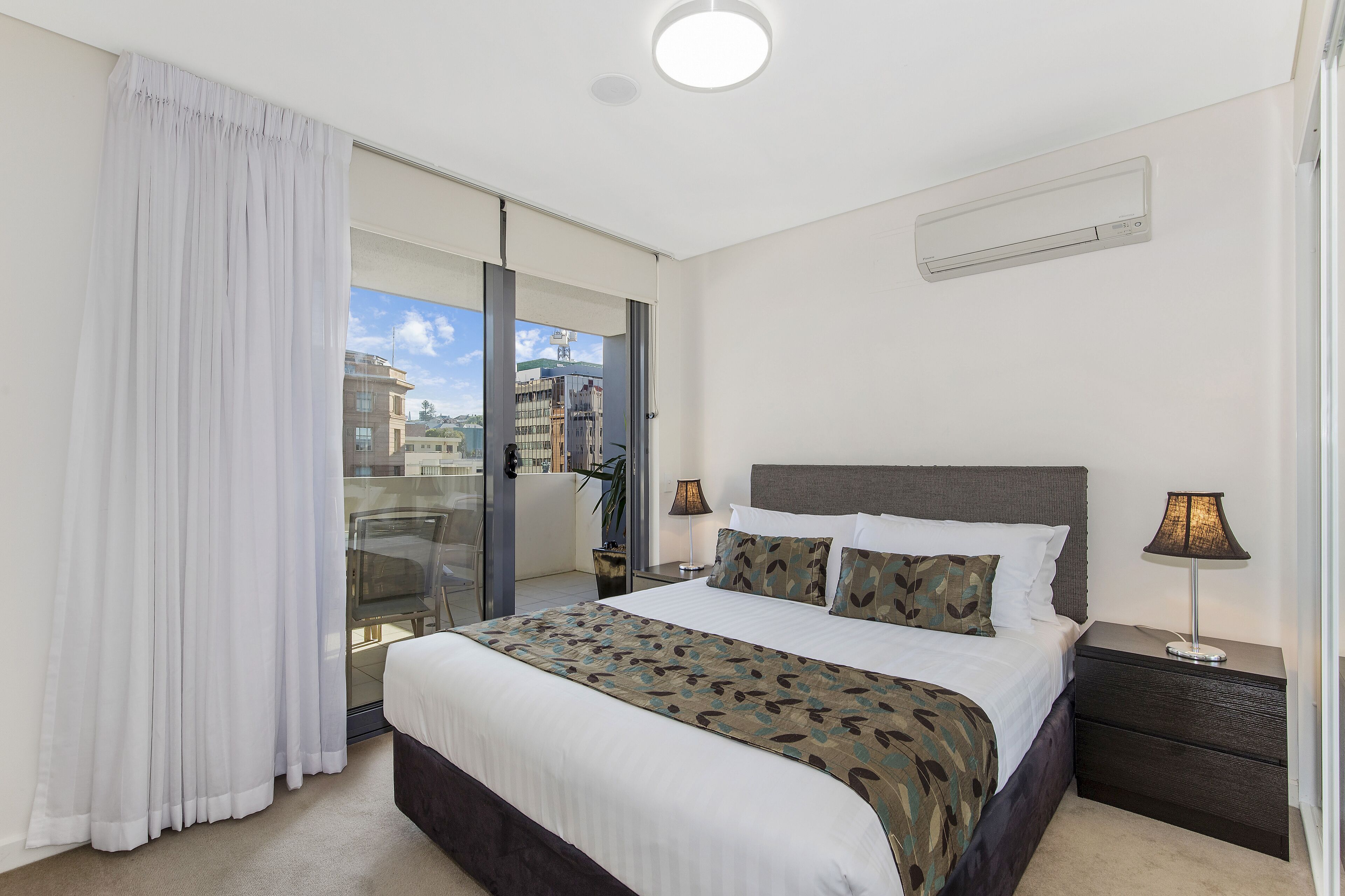 A modern hotel room with a large bed, a nightstand, a lamp, and a view through a glass door.