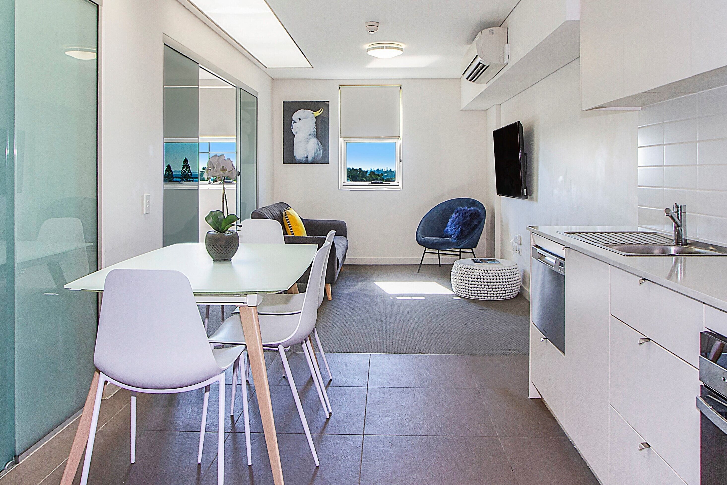 A modern kitchen with white cabinets, a dining area with a table and chairs, a blue chair, and a view of the outdoors through a window.