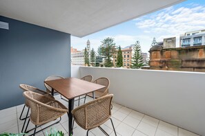 En balkong med ett bord och flätade stolar, med utsikt över en stadsmiljö med byggnader och träd.