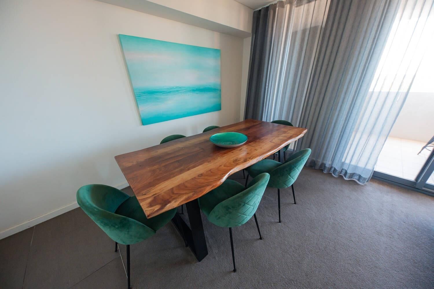 A dining area with a wooden table, green upholstered chairs, a teal painting on the wall, and a view through a window with curtains.