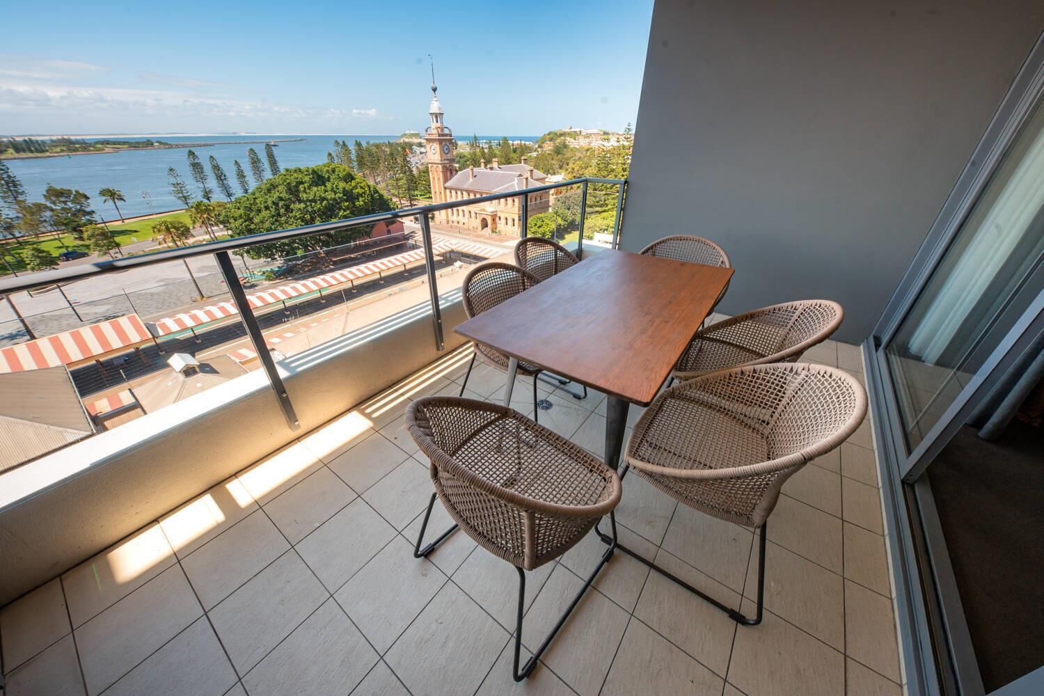 Un balcon avec une table et des chaises en osier, donnant sur un plan d’eau et un parc agrémenté d’un beffroi.