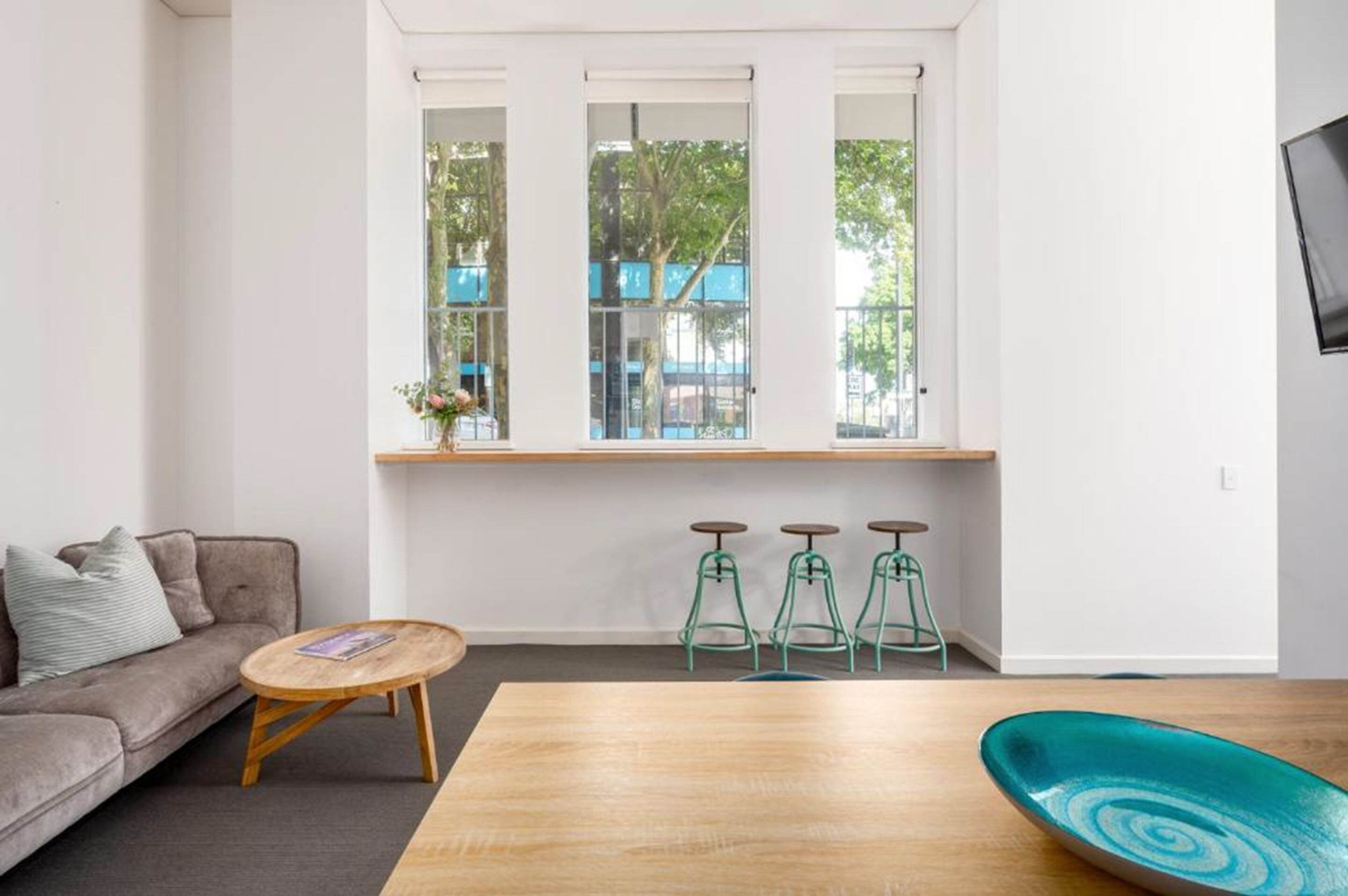 Een moderne woonkamer met een bank, een ronde houten tafel en drie groene barstoelen. Een groot raam biedt uitzicht op bomen en gebouwen.